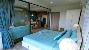 A bedroom with a large bed, a desk, and a chair. There is a kitchenette in the background. - Rain Cha-am Hua-hin by Noppawan (Cha-am)