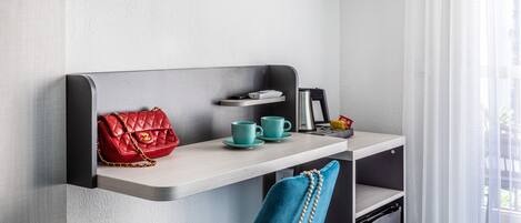 Una habitación de hotel con una cama, un escritorio con una silla, un televisor, una ventana con cortinas y un bolso rojo sobre el escritorio.