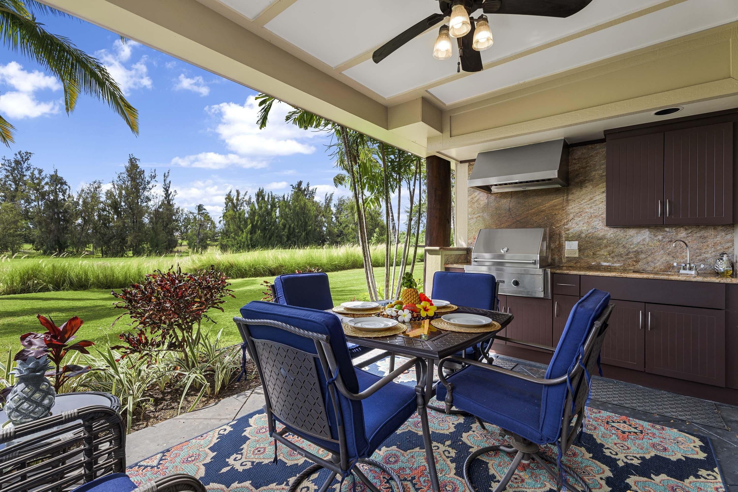 An outdoor dining area with blue chairs and a table set for two, overlooking a lush garden and a golf course.