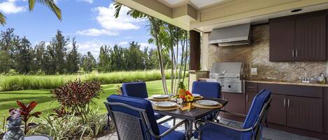 An outdoor dining area with blue chairs and a table set for two, overlooking a lush garden and a golf course.