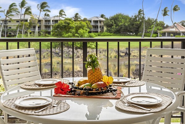 A round table set for four with white chairs, a tropical fruit centerpiece, and a view of a golf course and buildings in the background. - Shores At Waikoloa #234 At Bo Tree Tower 2 Bedroom Condo by RedAwning (Waikoloa)