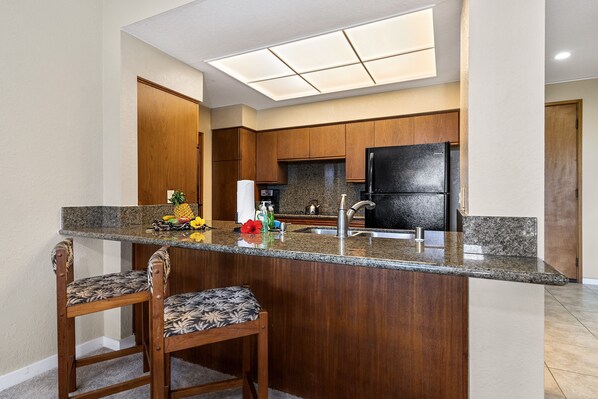 A kitchen with a granite countertop, wooden cabinets, a black refrigerator, and a small island with two bar stools.