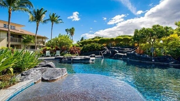 Une piscine agrémentée d’une cascade, entourée d’une végétation tropicale et bordée d’un bâtiment doté de balcons.