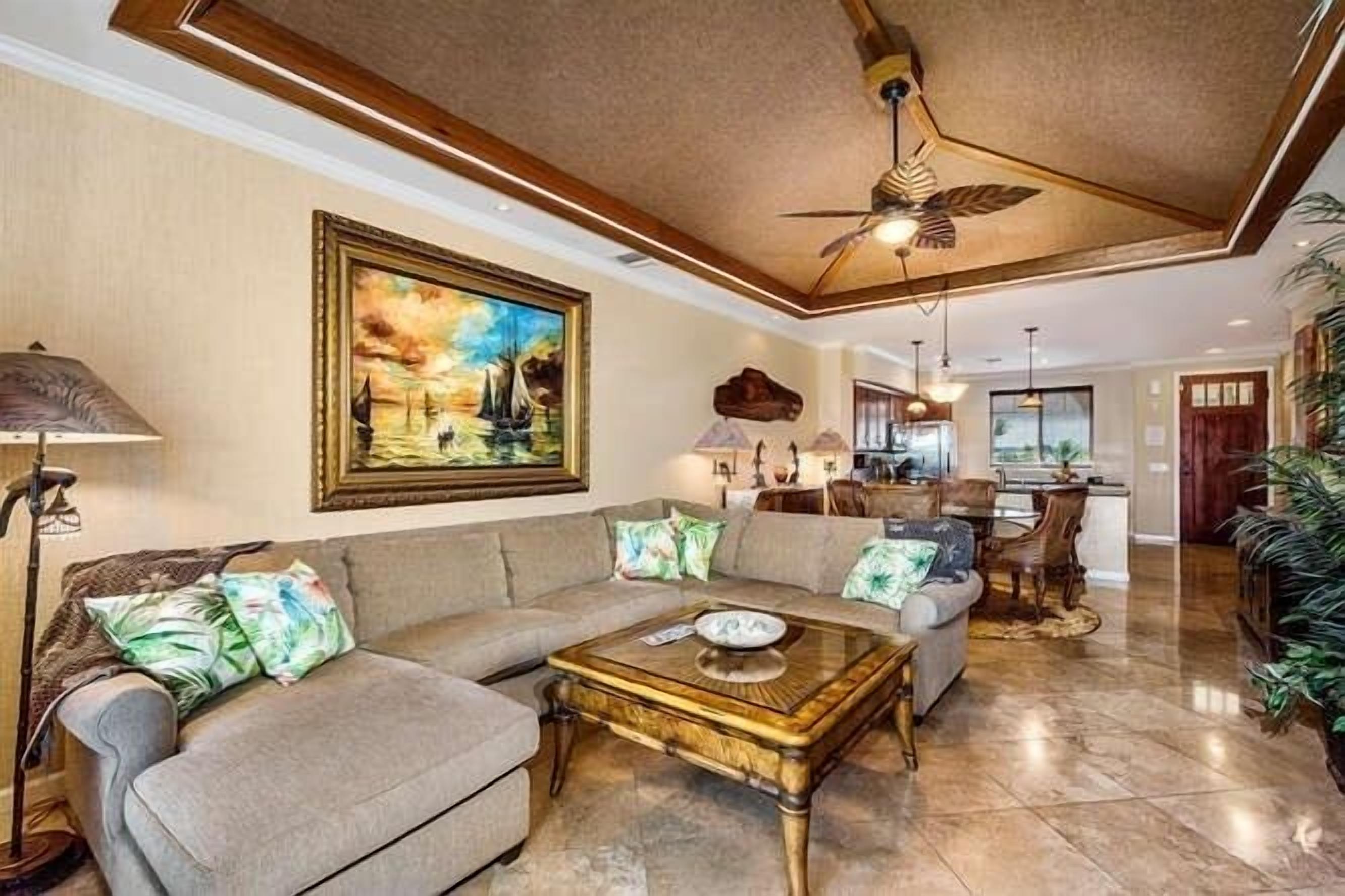 A spacious living room with a beige sofa, a wooden coffee table, and a ceiling fan. A painting is on the wall, and there is a kitchen area in the background.