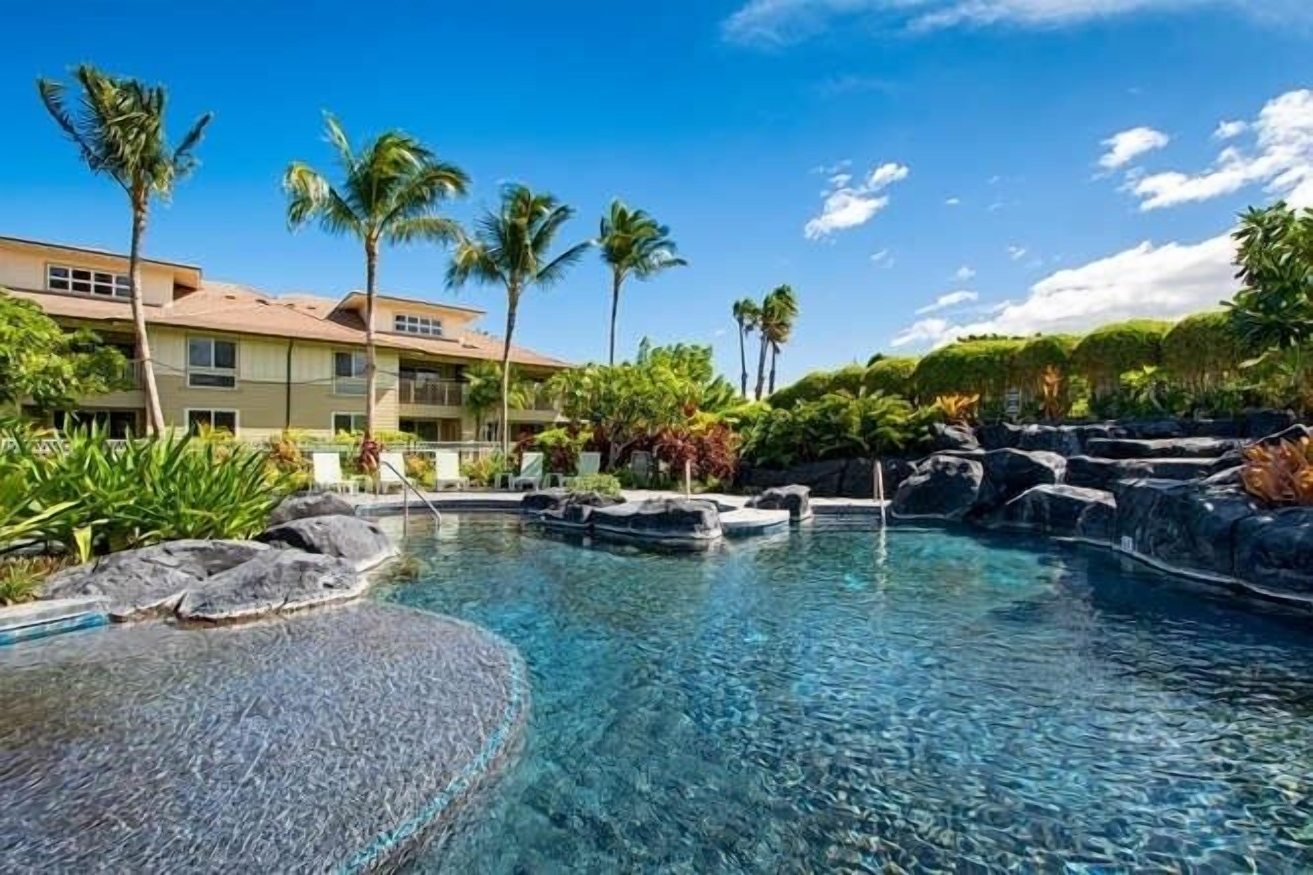 A hotel pool area with clear water, palm trees, and a building in the background.