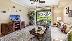 A living room with a television, a ceiling fan, a sofa, a coffee table, and a large window with a view of palm trees. - Waikoloa Beach S G2 2 Bedroom Condo by RedAwning (Waikoloa)