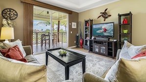 Ein Wohnzimmer mit Fernseher, Couchtisch und Sofa. Eine Glastür führt auf einen Balkon mit Gartenmöbeln.
