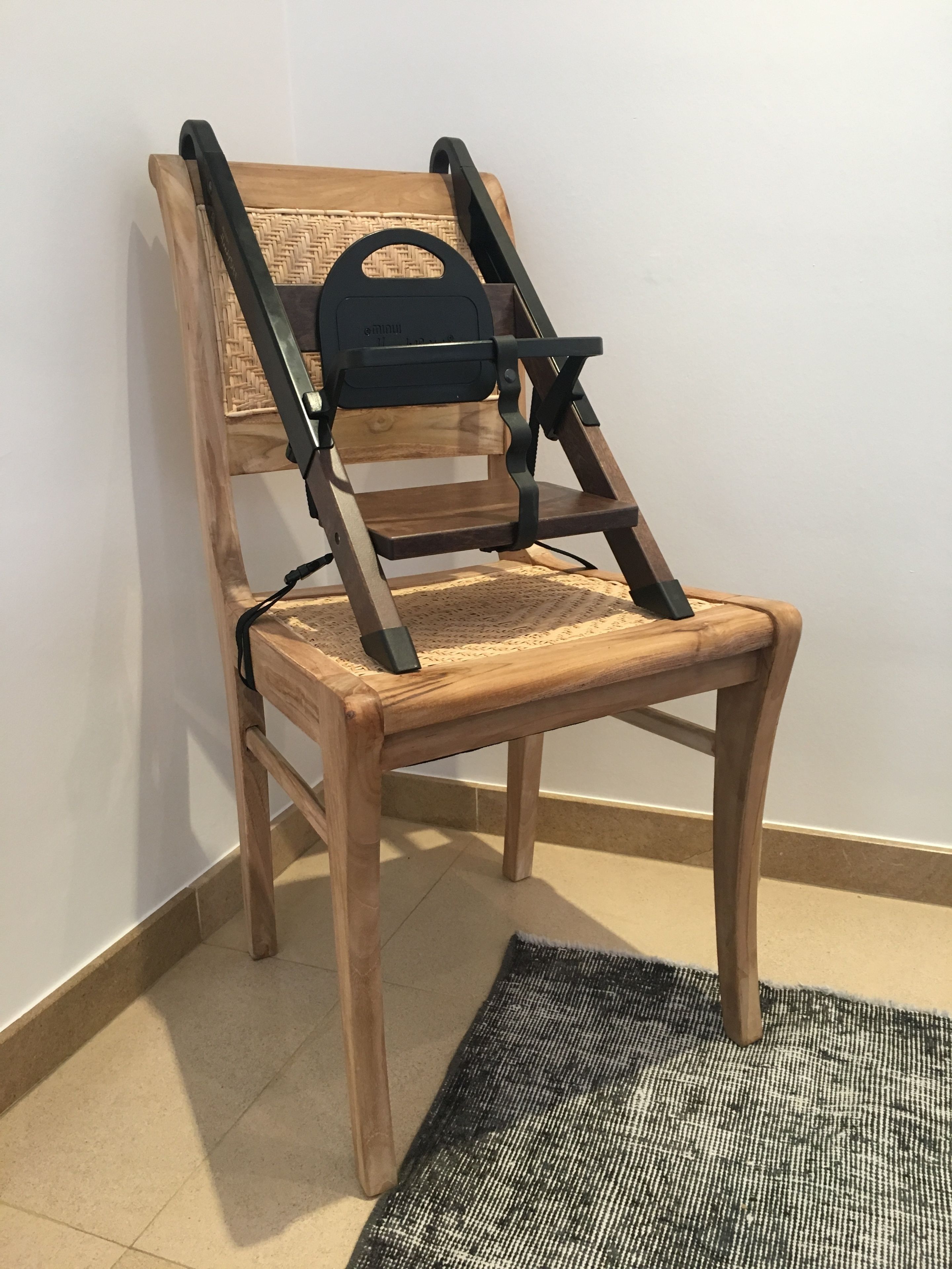 A wooden chair with a woven backrest and a black seat cushion, placed on a tiled floor with a black and white patterned rug.