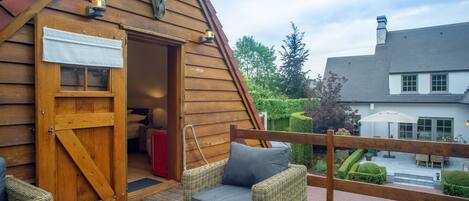 A wooden cabin with a large open door, wicker furniture, and a view of a garden and neighboring buildings.