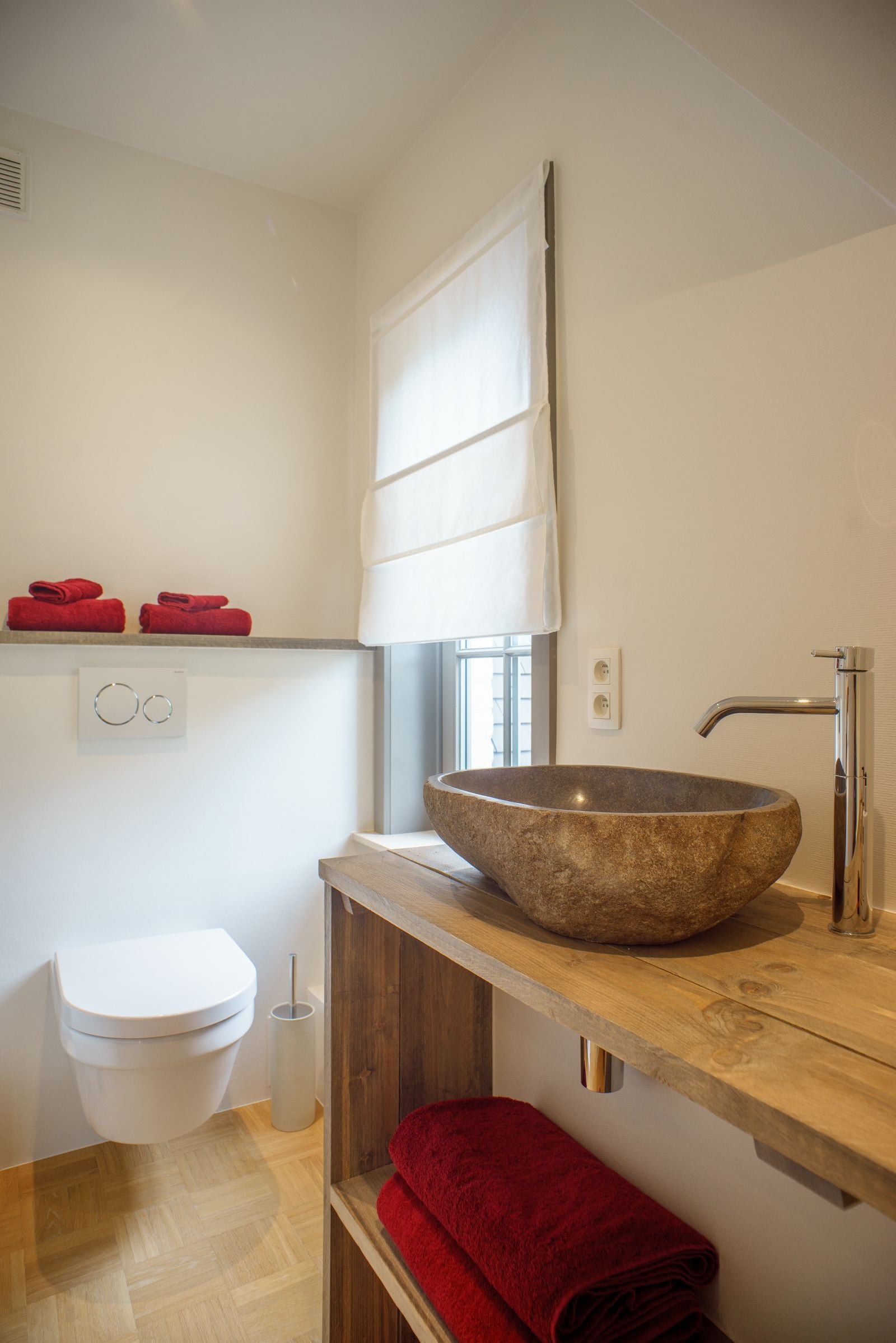 Un baño moderno con lavamanos de piedra, inodoro y estante de madera con toallas rojas.