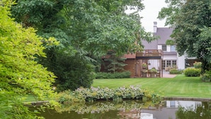 A serene garden with a pond, lush greenery, and a house with a wooden deck and outdoor furniture. - Villa Magdalena (Eeklo)