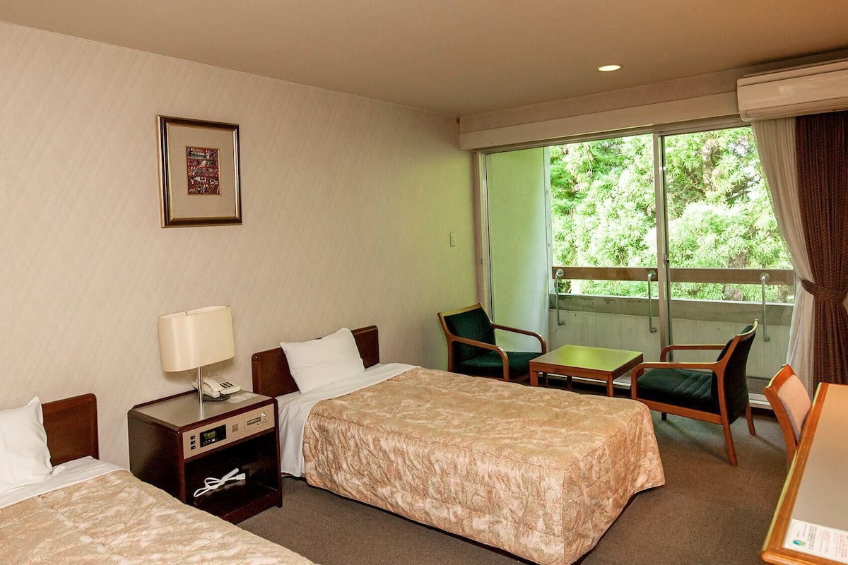 Une chambre d’hôtel avec deux lits, un bureau, une chaise, un téléphone et une fenêtre donnant sur un espace verdoyant.
