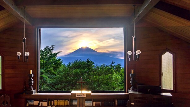A room with a large window providing a view of a mountain and trees. There are bookshelves and a cross on the windowsill. - YMCA International Youth Center TOZANSO (Gotemba)