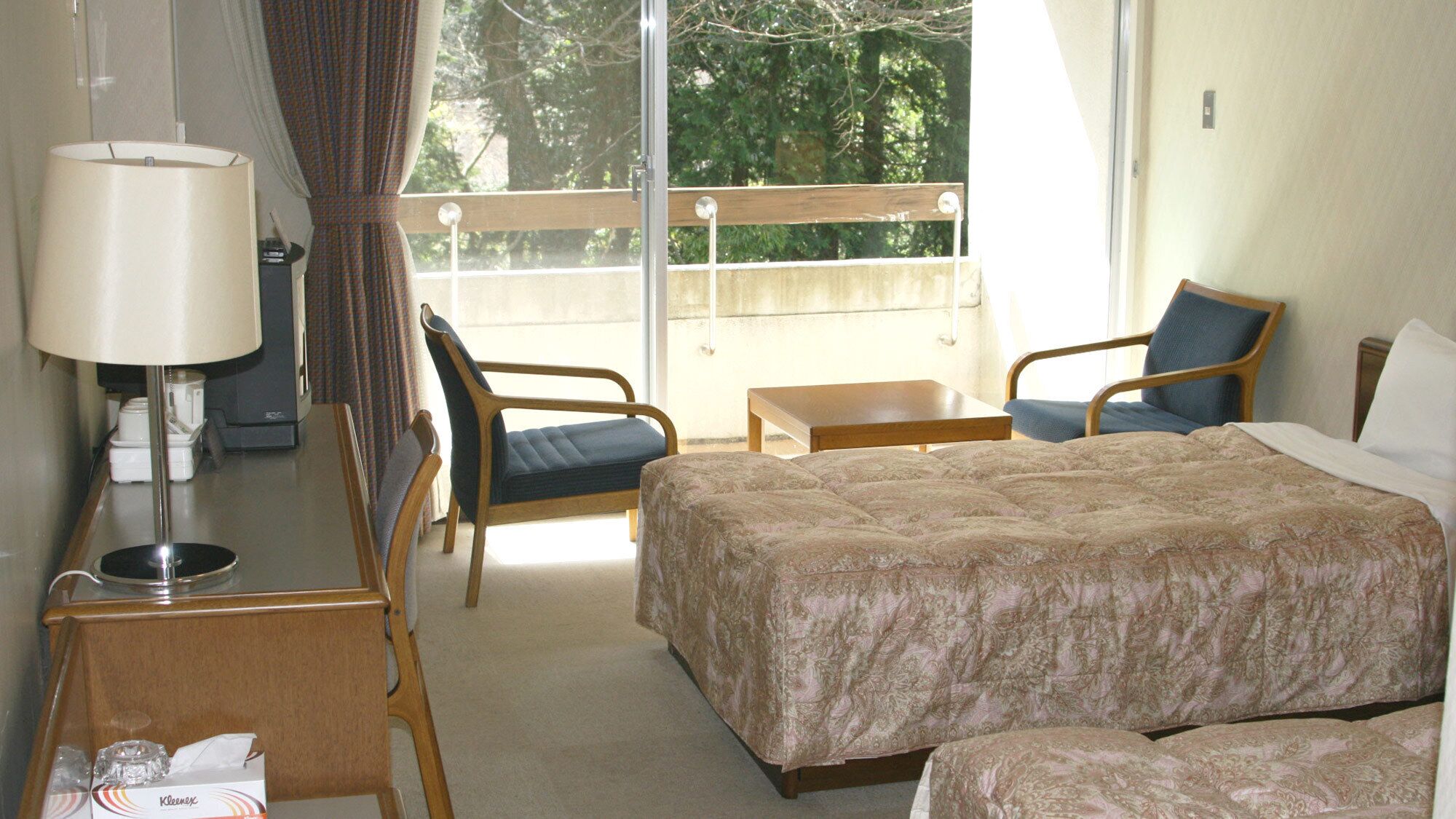 Une chambre d’hôtel avec un lit, deux chaises, une petite table, une lampe et une vue sur les arbres par la fenêtre.