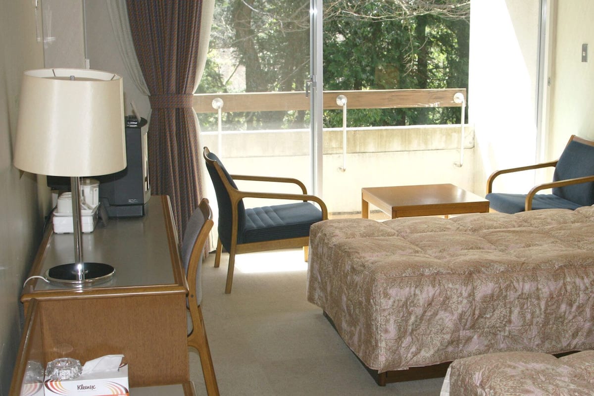 Une chambre d’hôtel avec un lit, deux chaises, une petite table, une lampe et une vue sur les arbres par la fenêtre.