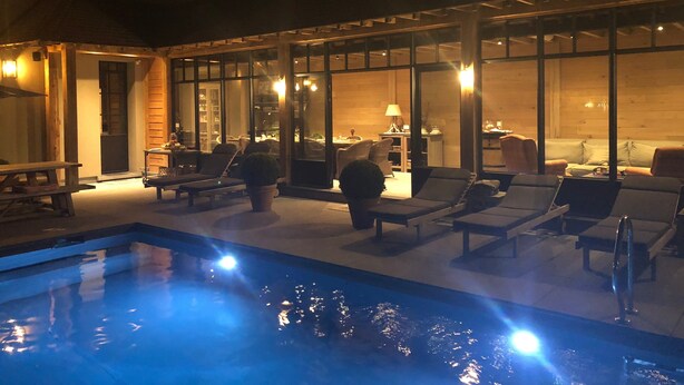 Zona de piscina exterior con sillones de descanso y un área de estar, rodeada de paredes de vidrio y vigas de madera.