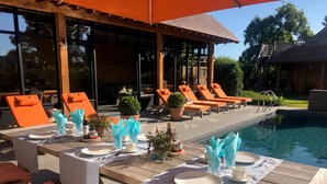 Un área de comedor junto a la piscina con mesas de madera, sillones reclinables naranja y una piscina.