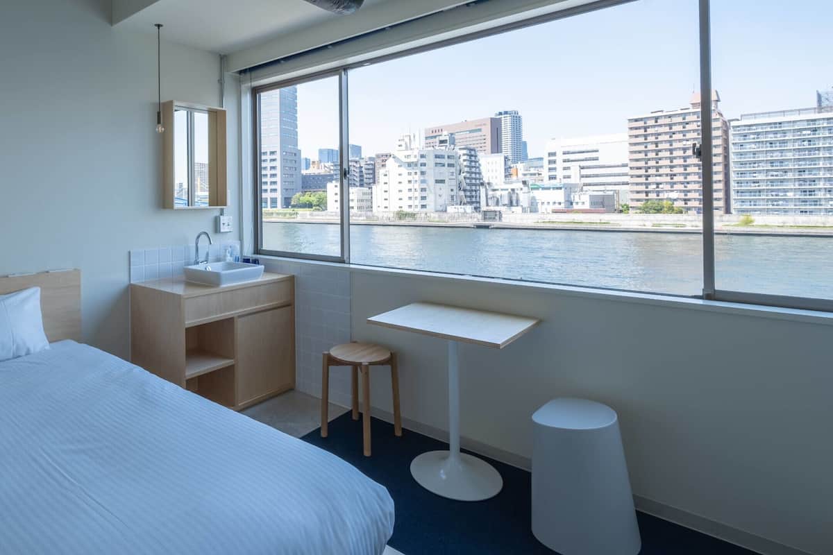 A hotel room with a bed, a small desk, a sink, and a view of a cityscape through large windows.