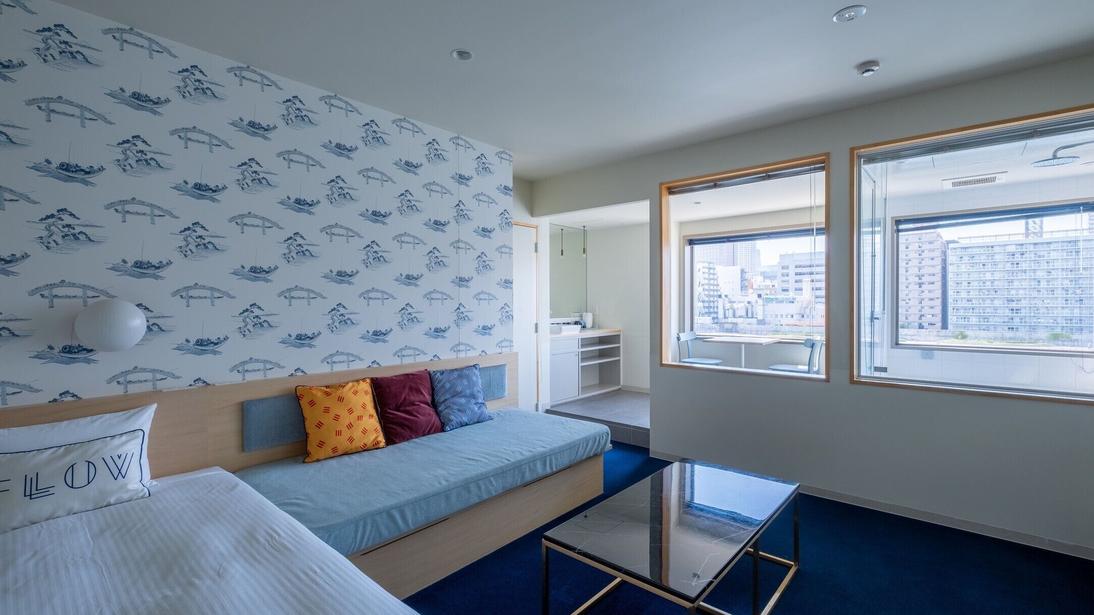 Quarto de hotel moderno com um sofá azul, uma mesa de centro de vidro e vista para a cidade através de grandes janelas.