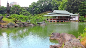 Ein traditionelles japanisches Gebäude mit einem überdachten Pavillon an einem Teich, umgeben von Steinen und Grünpflanzen.