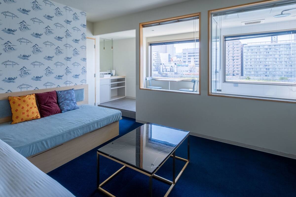 A modern hotel room with a bed, a sofa, a glass coffee table, and a view of the city through large windows.