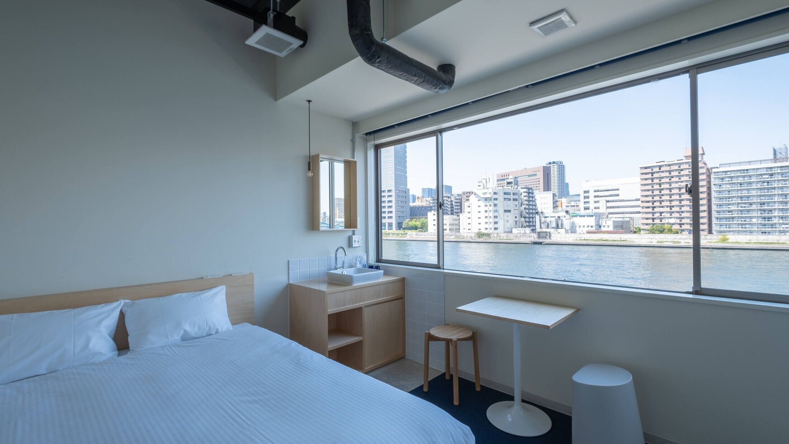 Quarto de hotel com cama, uma pequena escrivaninha, espelho e janela com vista para a cidade.