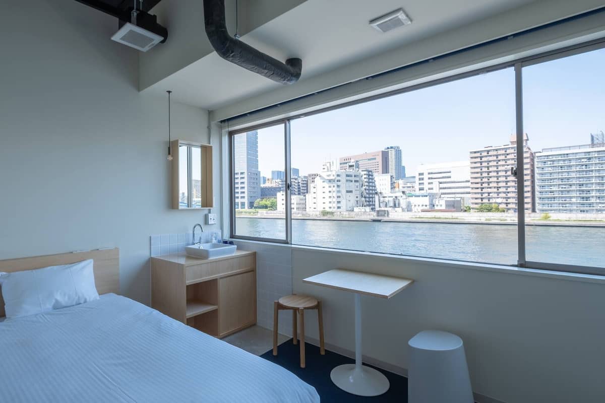 A hotel room with a bed, a small desk, a mirror, and a window with a city view.