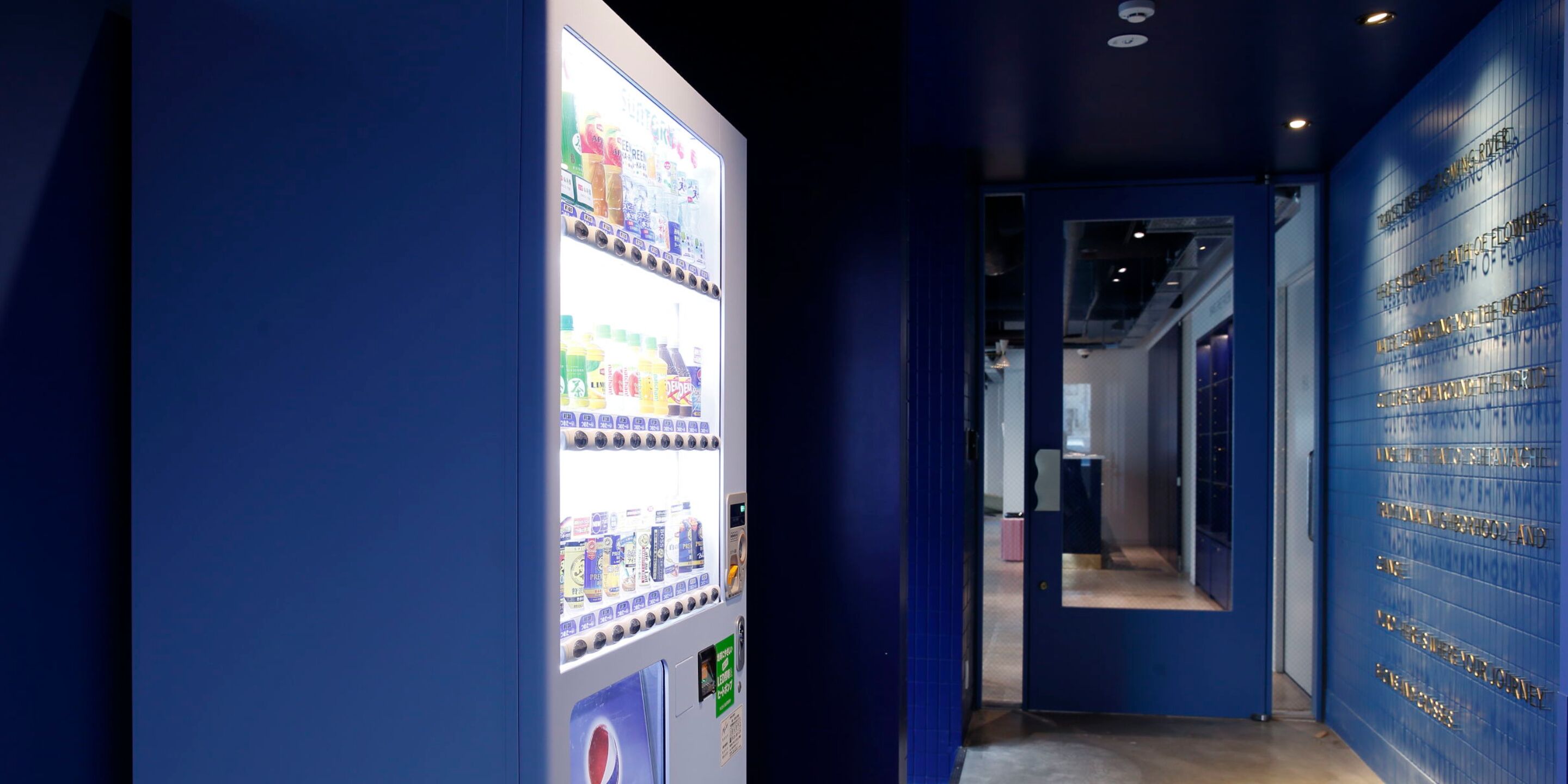 A hallway with a vending machine stocked with beverages and snacks, a blue wall with engraved text, and a door leading to another room.