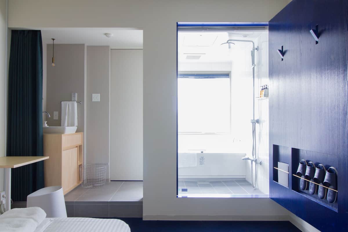 A hotel room with a bed, a small table, a mirror, a window, and a shower area.