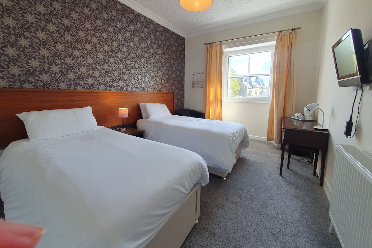A hotel room with two beds, a wooden headboard, a nightstand with a lamp, a desk with a computer, a window with curtains, and a television on the wall.