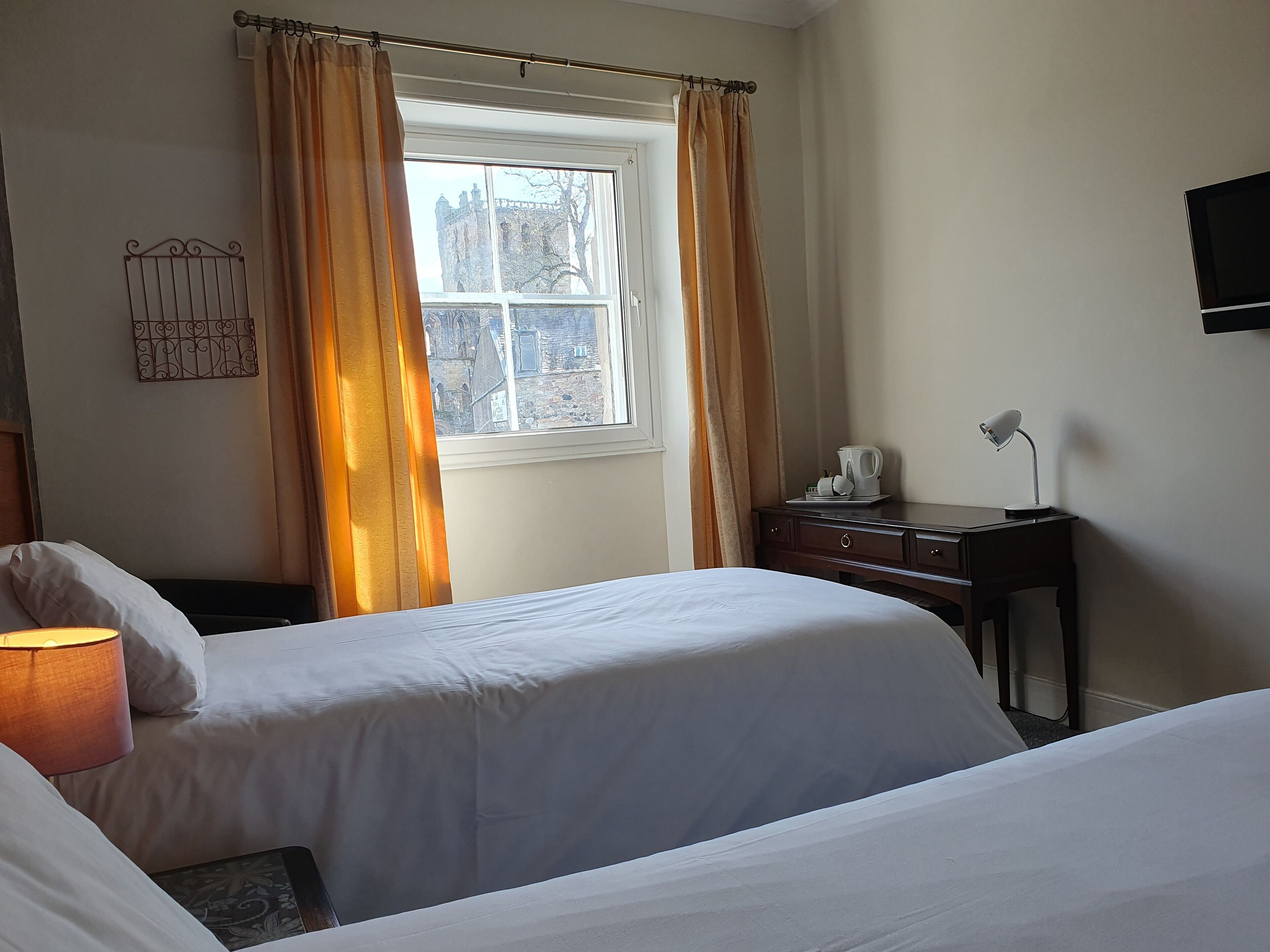 A hotel room with two beds, a window with curtains, a bedside table with a lamp, and a wall-mounted TV.