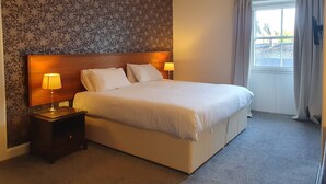 A neatly made bed with two pillows and two bedside lamps, a wooden headboard, and a window with curtains. - The Royal Hotel (Jedburgh)