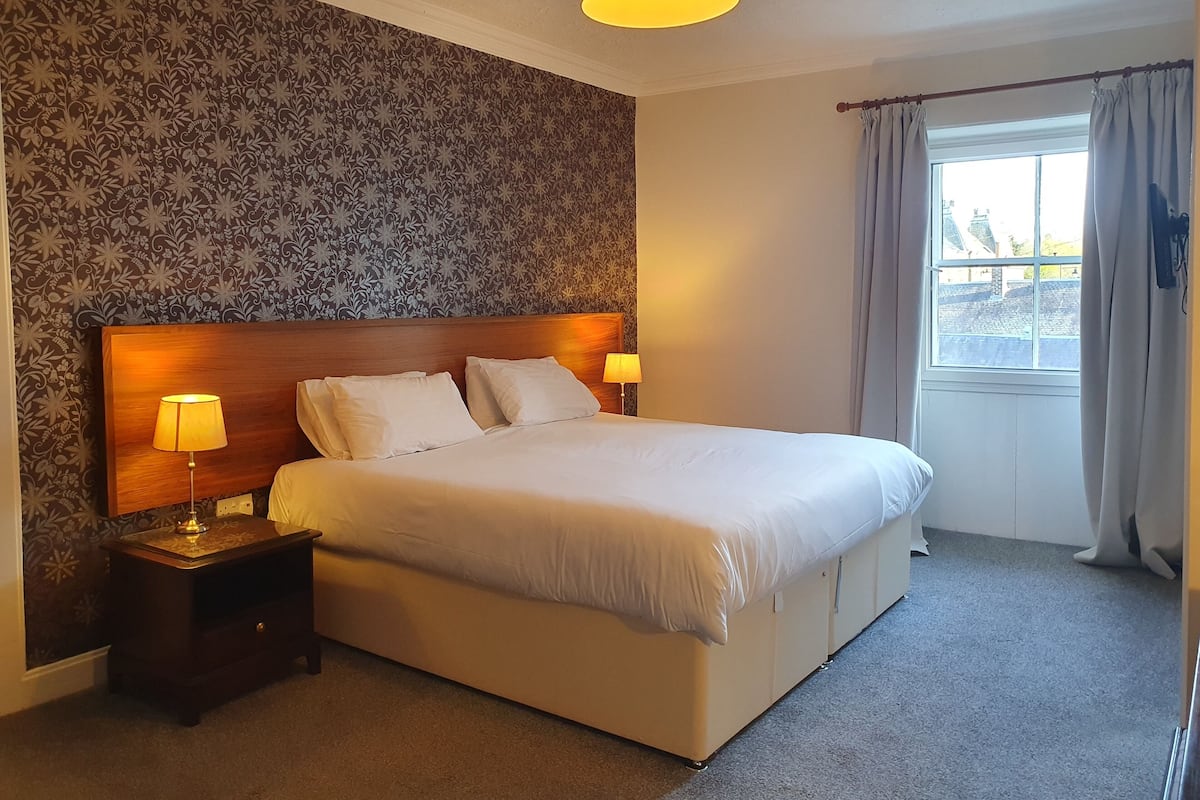 A neatly made bed with two pillows and two bedside lamps, a wooden headboard, and a window with curtains.