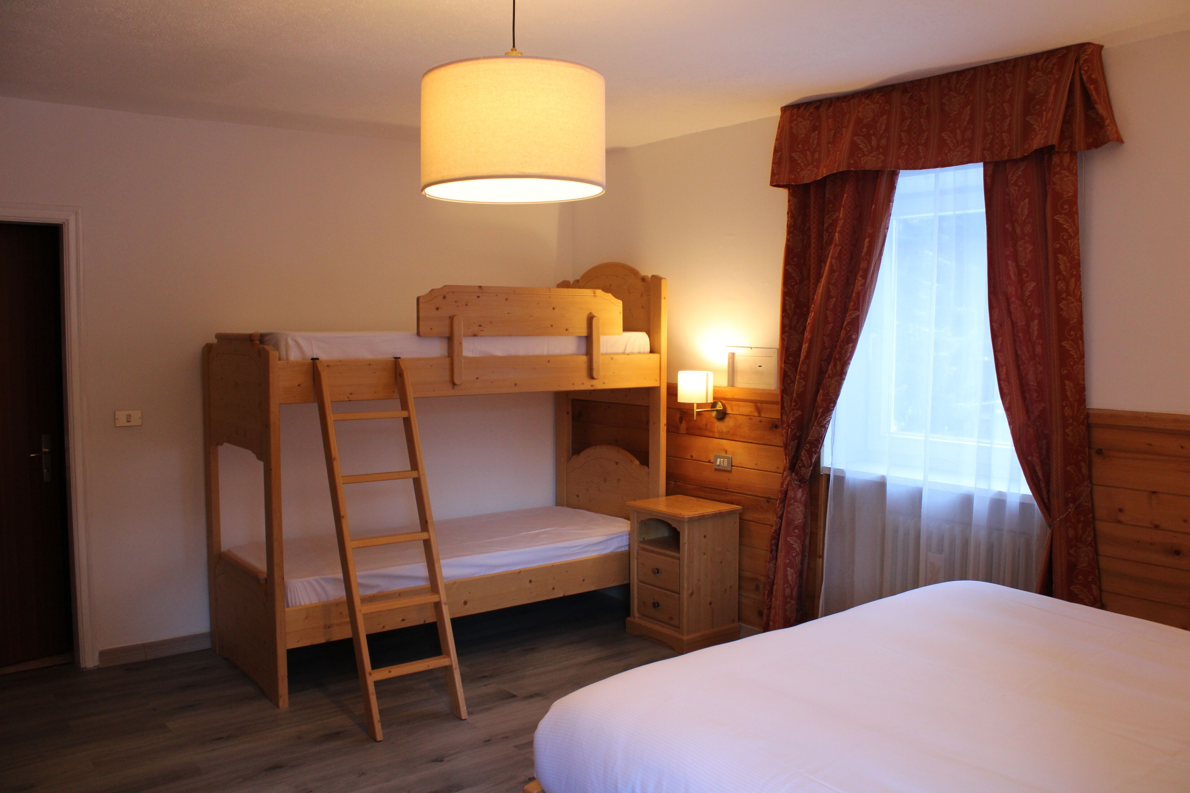 A room with a bunk bed, a desk with drawers, a small bedside table with a lamp, and a window with curtains.