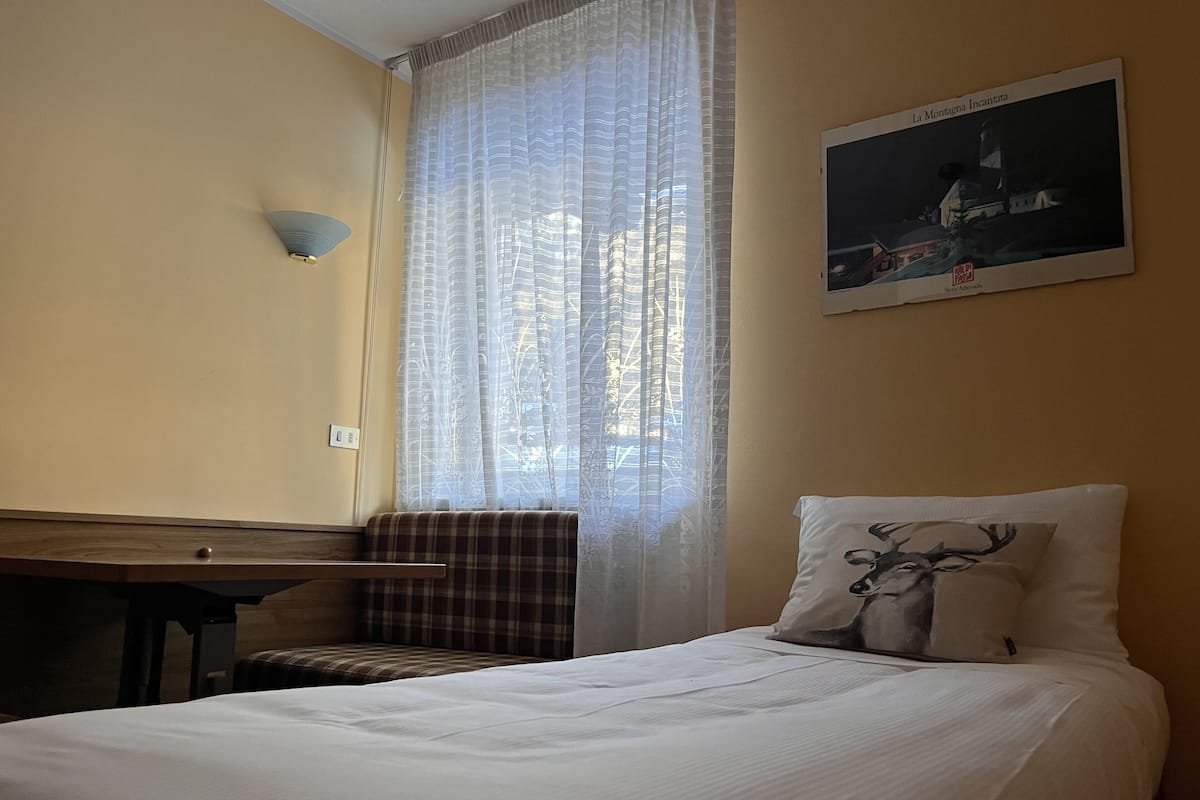 A hotel room with a bed, a chair, a desk, and a window with sheer curtains.