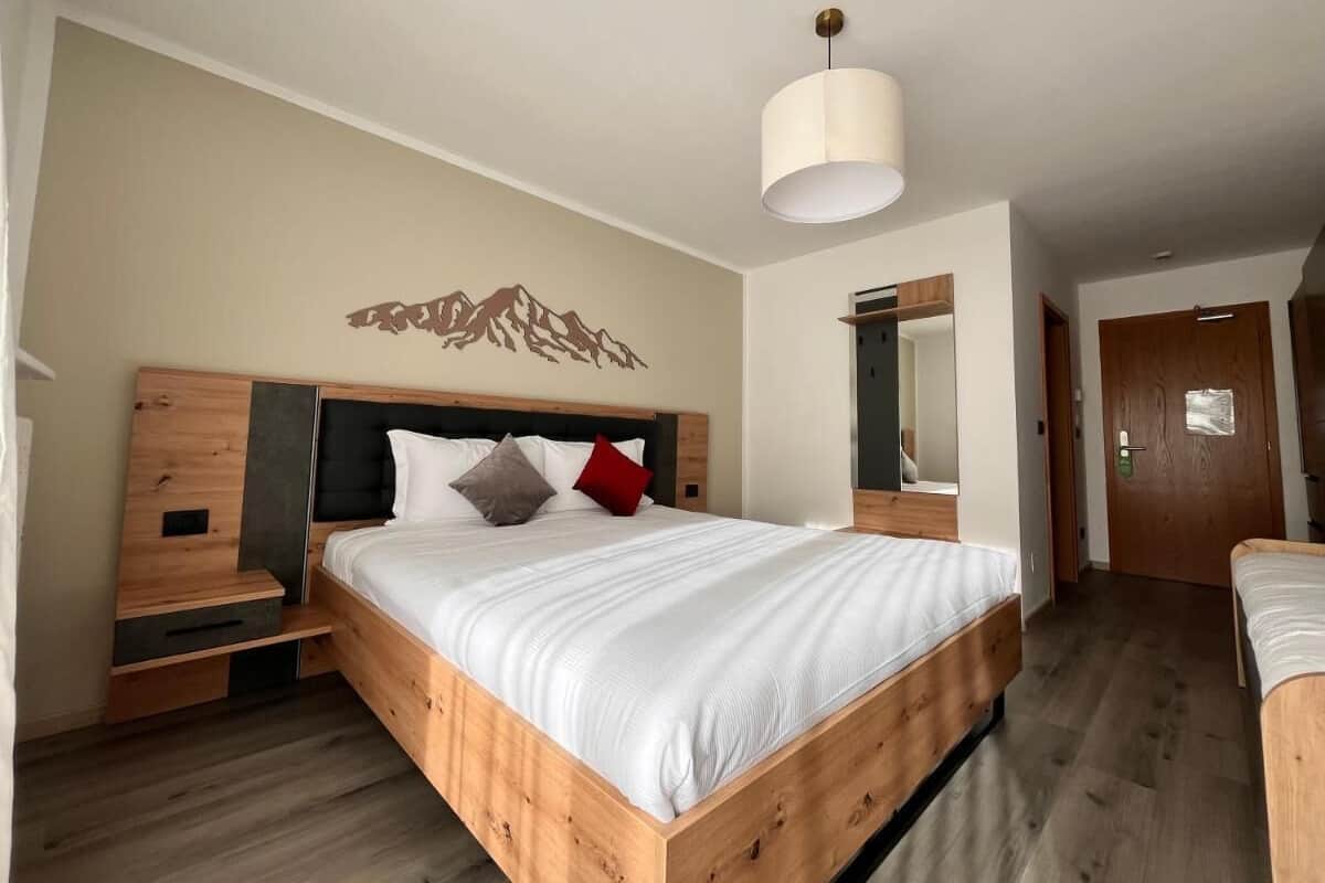 A modern bedroom with a wooden bed, a nightstand, a bedside table, a wall-mounted mirror, and a door.