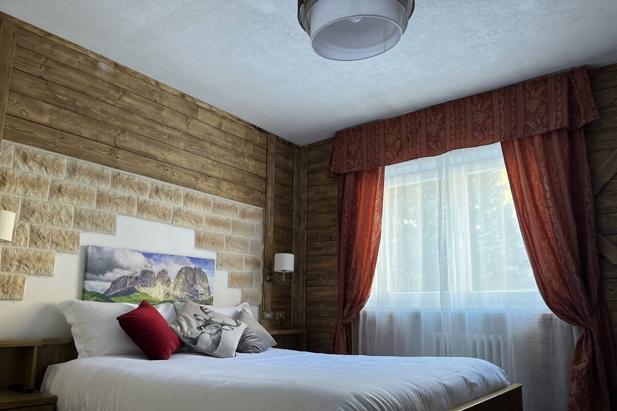 A bedroom with a wooden wall, a bed with white bedding, a red pillow, and a painting of mountains.