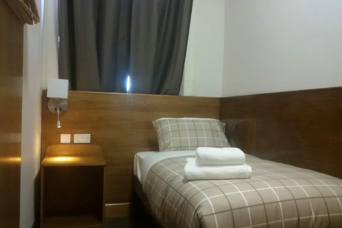A hotel room with a wooden headboard, a single bed with a checkered bedspread, a pillow, a nightstand with a lamp, and a window with curtains.