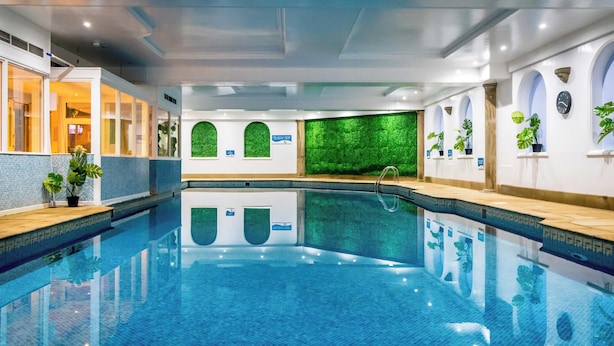 A large indoor swimming pool with clear blue water, surrounded by walls with green textured backdrops and potted plants. - The Sleepwell Inn (Workington)