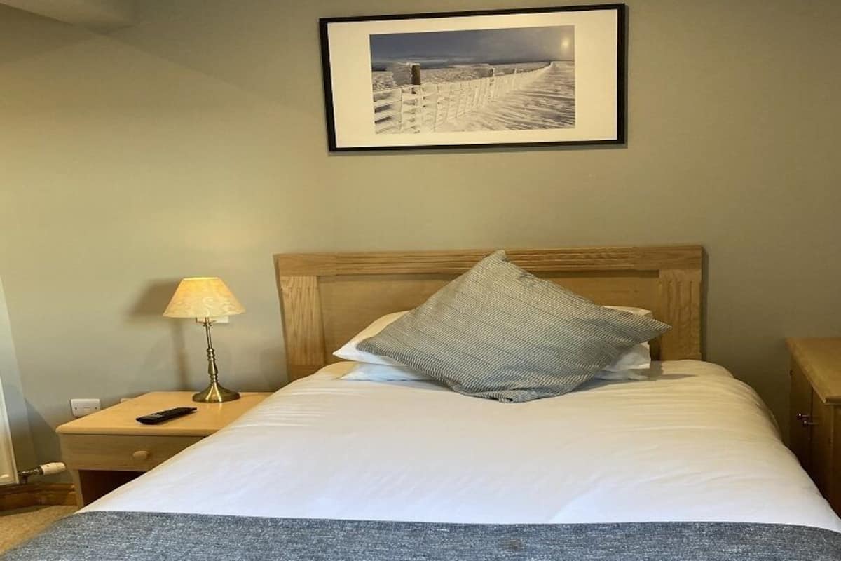 Una cama bien hecha con cabecera de madera, una lámpara de noche y un cuadro en la pared.