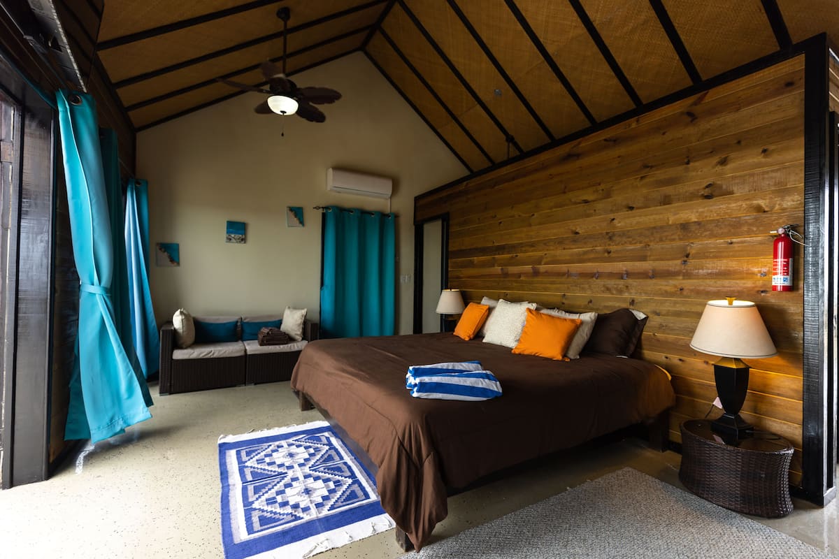 A bedroom with a wooden ceiling, a bed with brown bedding, a blue and white patterned rug, a wicker side table with a lamp, and a ceiling fan.