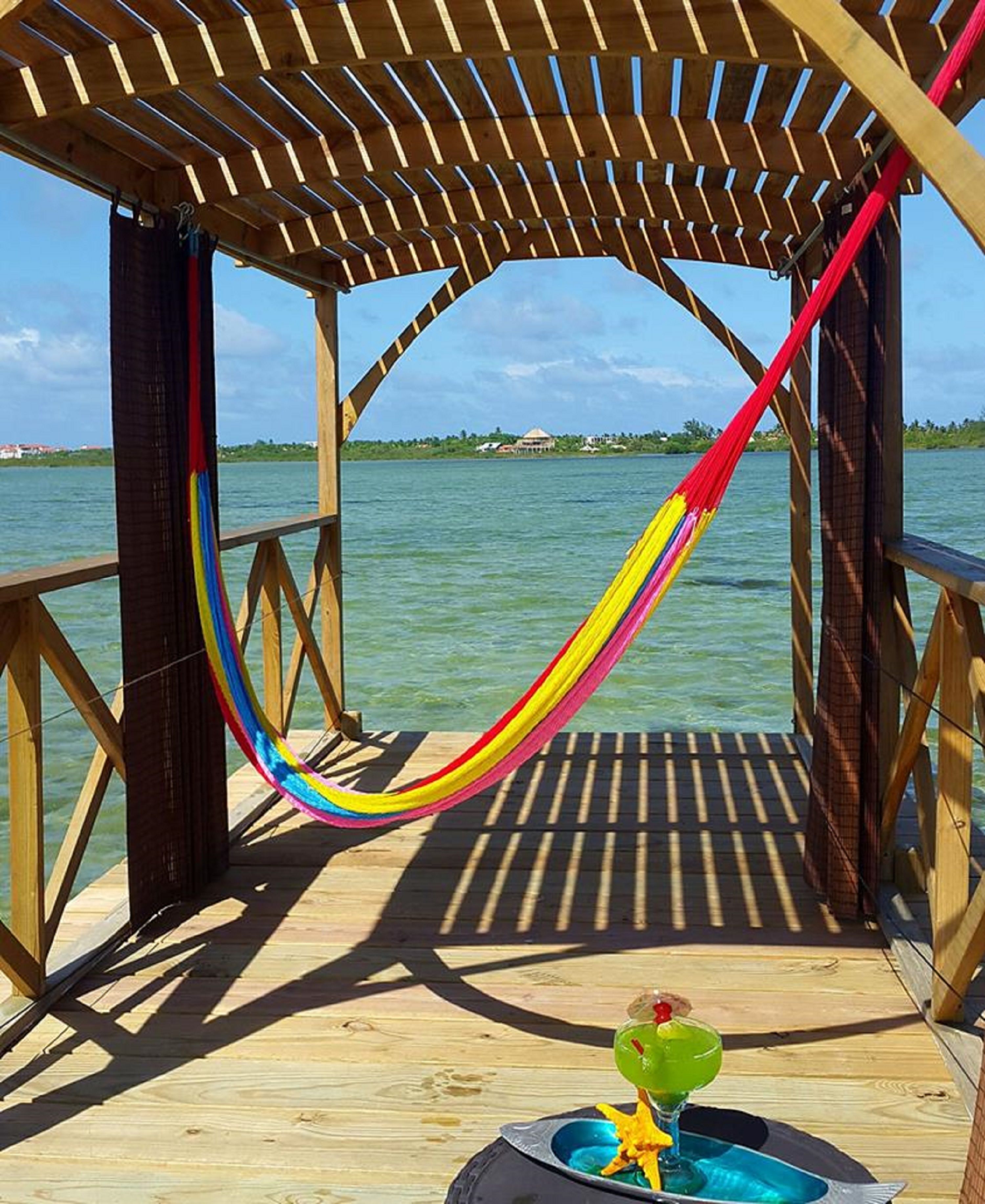 A wooden deck with a hammock and a drink on a table, overlooking a body of water.