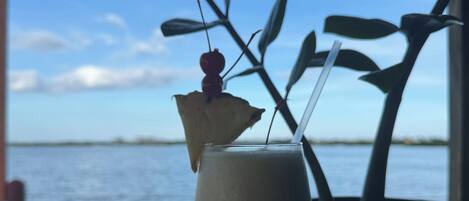 Une boisson, agrémentée d’une garniture, posée sur une table près d’une fenêtre donnant sur un plan d’eau.