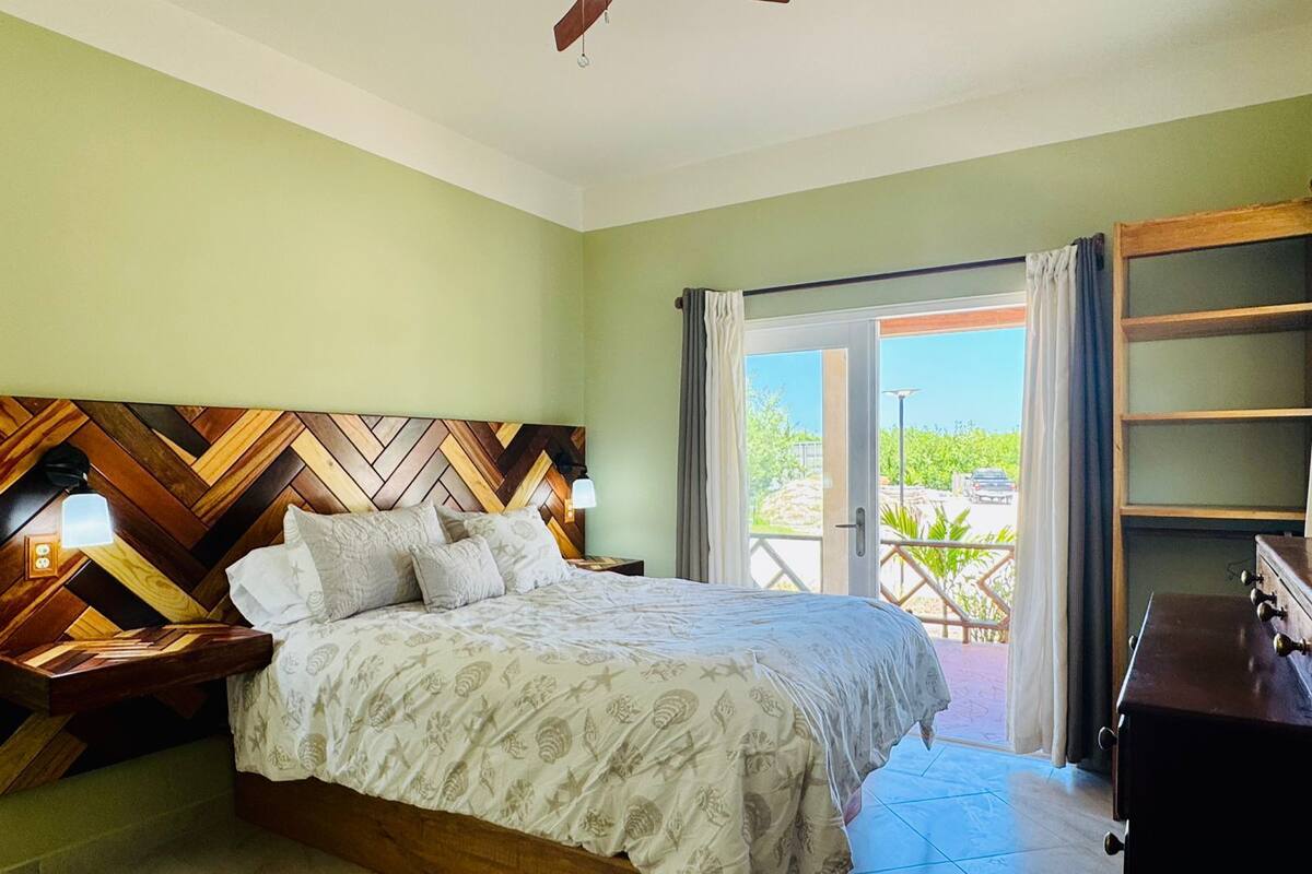 A bedroom with a bed, wooden headboard, nightstands, a dresser, and a balcony with a view.
