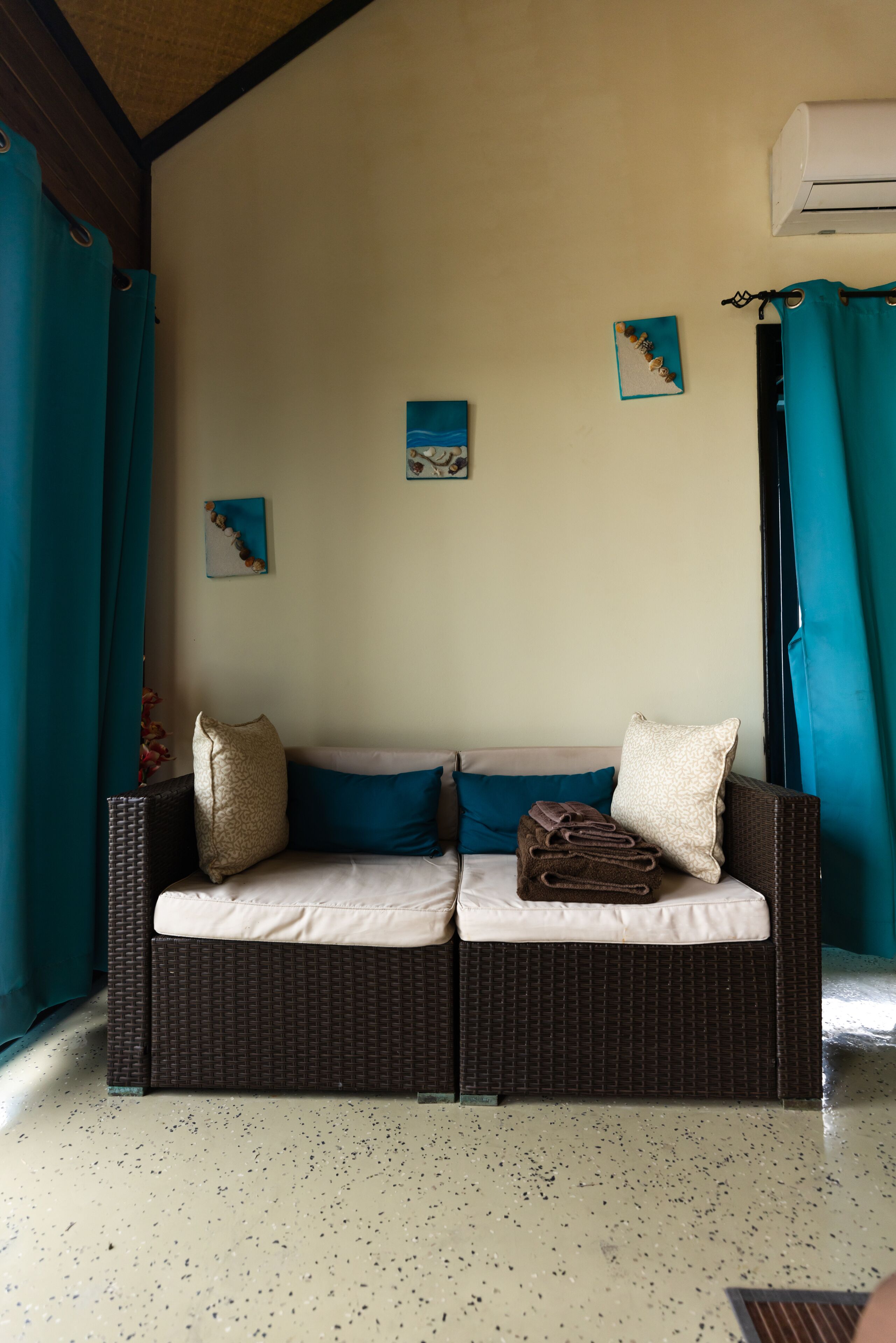 A room with a brown wicker sofa, teal curtains, and two wall-mounted paintings.
