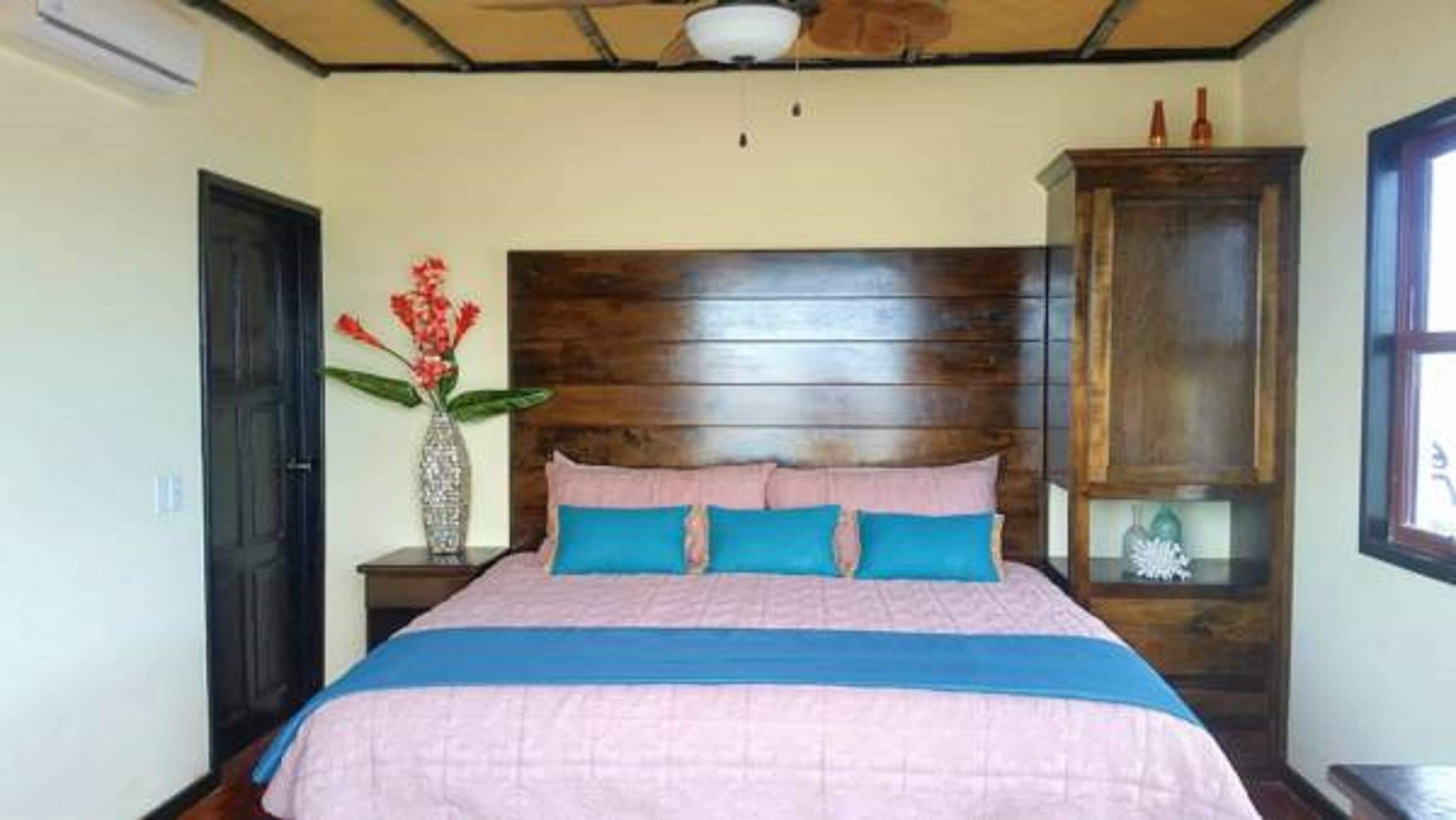 A bedroom with a wooden headboard, a bed with pink and blue bedding, a vase with flowers, and a cabinet.