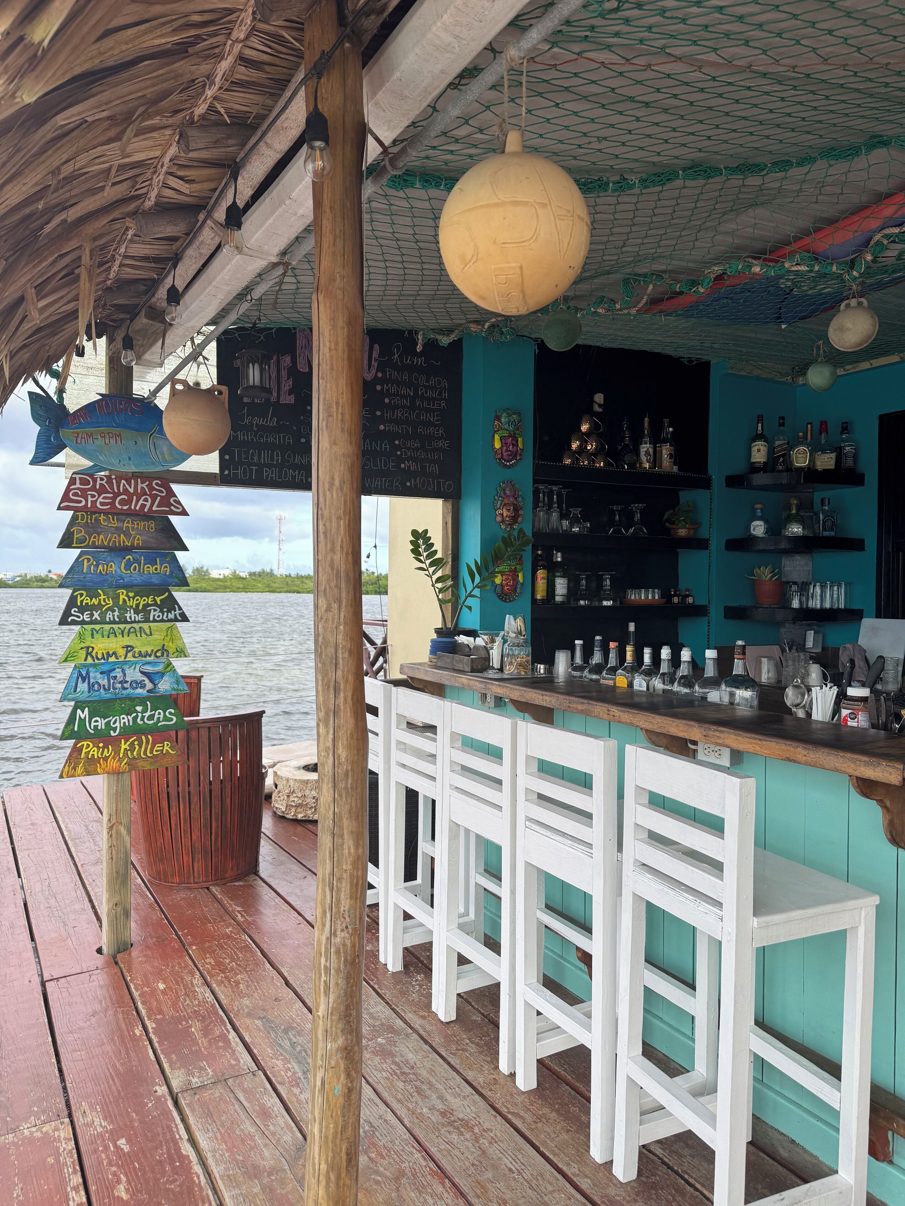 A bar with a wooden counter and white barstools, a sign listing drinks, and a view of the water.