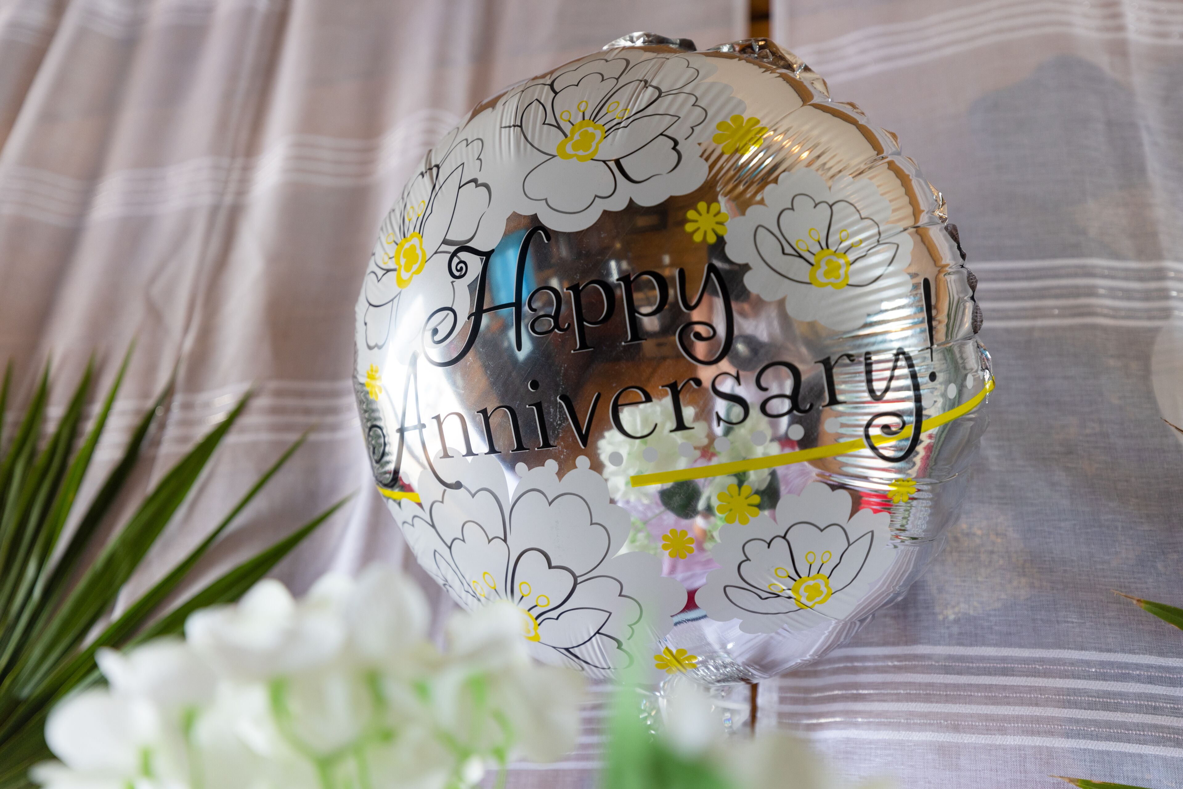 Balão prateado com a frase 'Happy Anniversary' escrito, colocado sobre uma superfície com padrão listrado.
