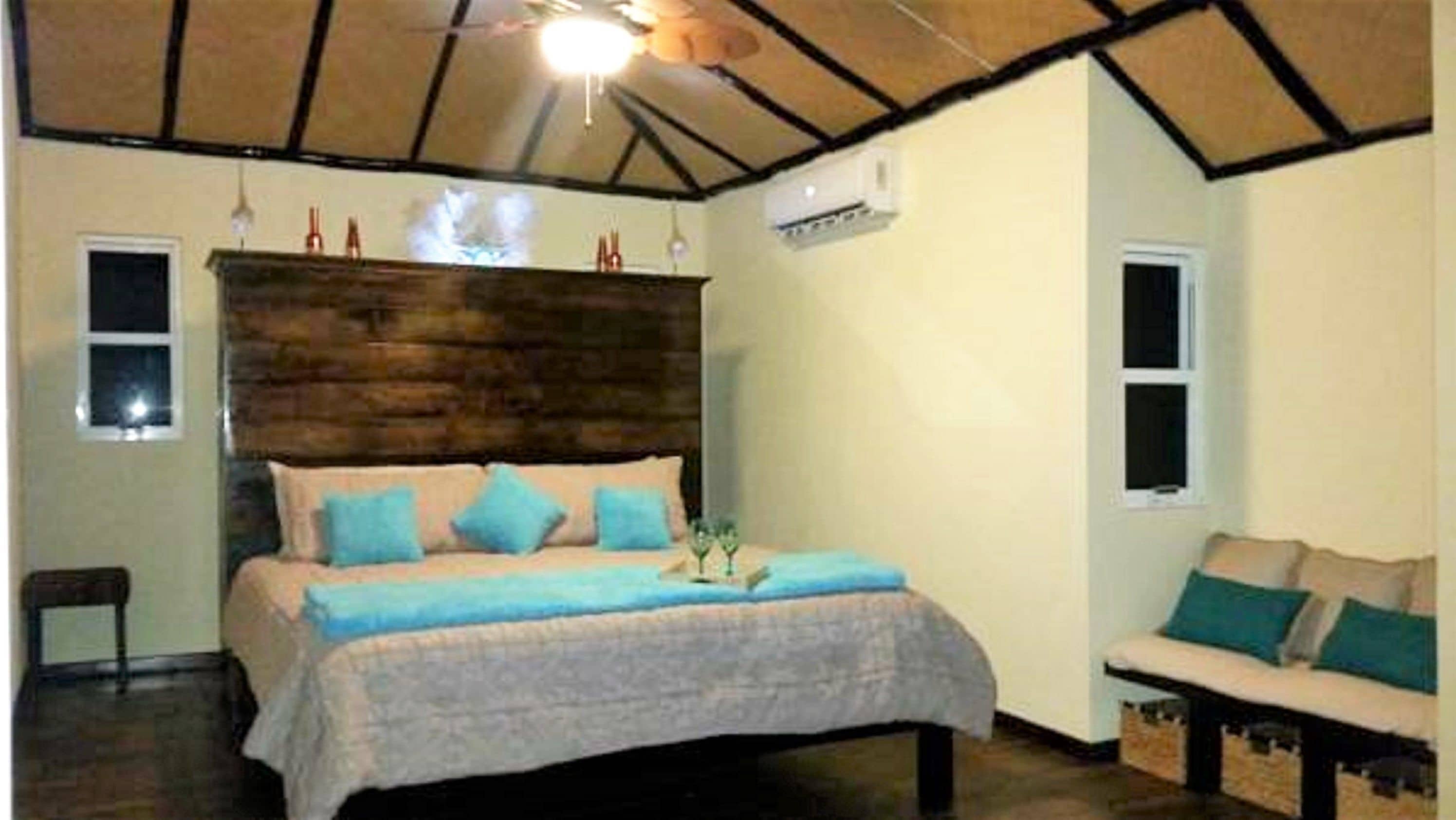 Un dormitorio con cabecera de madera, cama con ropa de cama azul, mesita de noche y un banco con cojines.