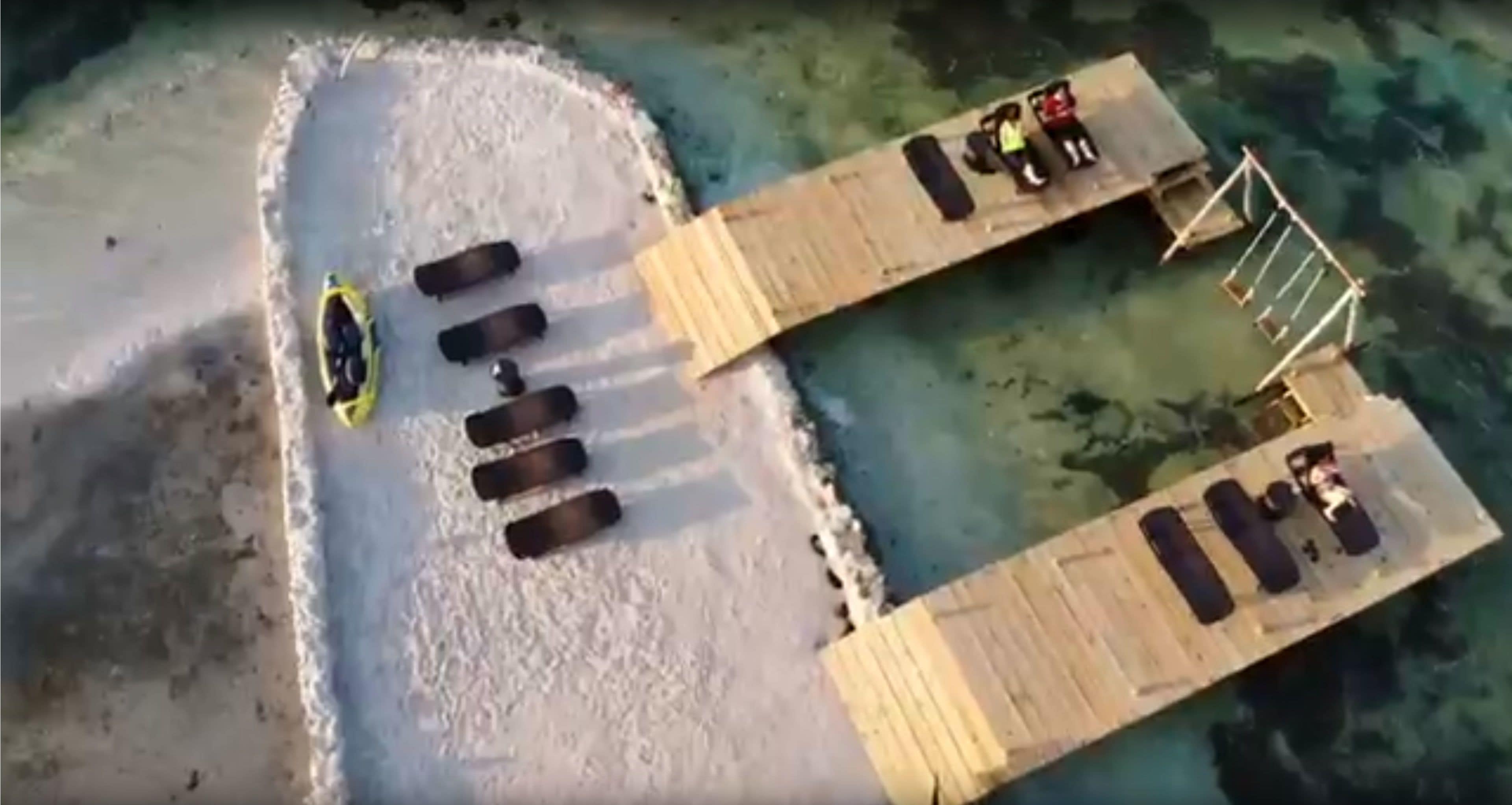 An aerial view of a beach with a wooden pier extending into the water, featuring several lounge chairs and a yellow kayak.